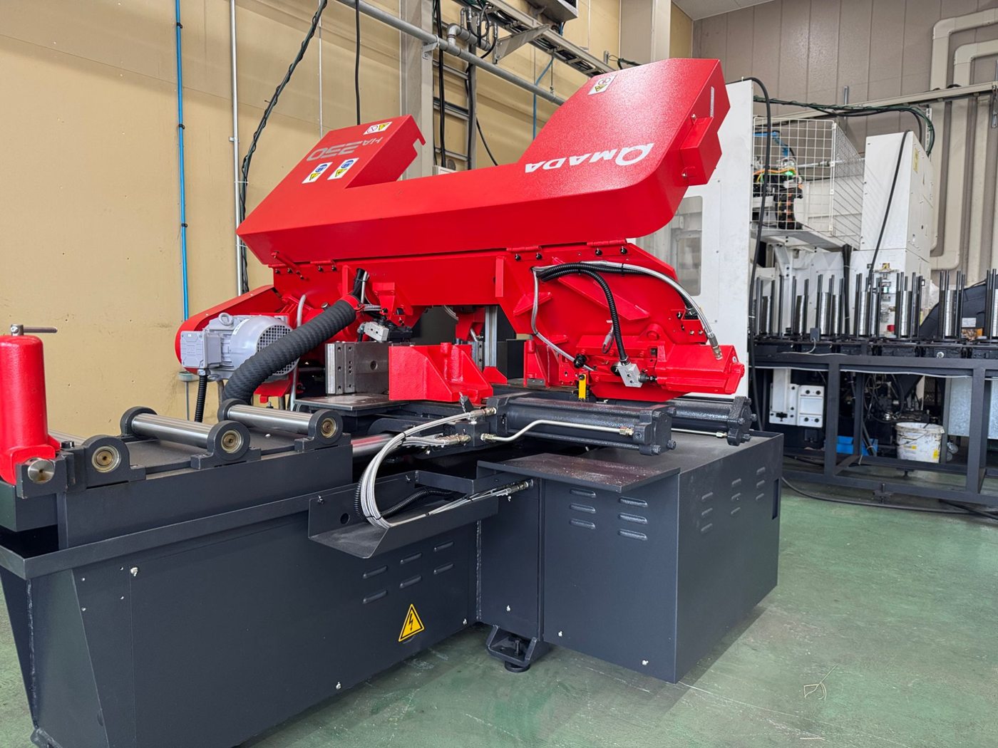 AMADA のバンドソー切断機 HA250。赤い鋸のヘッドと、鋼材を送るローラー、材料を押さえるバイス